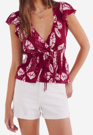 Vrouw draagt een rood-wit patroon wikkelstijl top met vlindermouwen en witte shorts met opgerolde zoom, poseert met één hand op de heup.