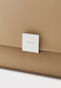 Hnedá kožená taška s textúrou, s strieborným obdĺžnikovým zapínaním a logom "Calvin Klein". Hladký povrch, štruktúrovaný tvar, minimalistický dizajn.