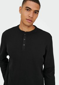 Sort langærmet henley skjorte med en tekstureret stof, der har en rund halsudskæring og en stolpe med tre sorte knapper ved brystet.