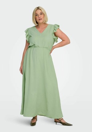 Žena v světle zelených, bezrukávých, na kotníky dlouhých šatech s volánkovými rameny a olivově zelených botách s špičatou špičkou, stojící s jednou rukou na boku.