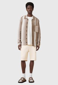 Camicia a righe con bottoni in beige e marrone con taschino. Indossata sopra una maglietta bianca, abbinata a pantaloni corti beige e sandali marroni.