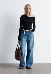 Schwarzes geripptes Oberteil mit schulterfreiem Schnitt, hochgeschnittene blaue weite Jeans, braune Wildleder-Tasche und braune Schuhe mit strukturierten Details.