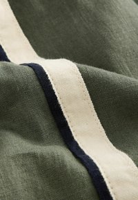 Olive žalia tekstilė, puošta šviesiai kreminėmis ir tamsiai mėlynomis juostomis. Audinys turi minkštą, tekstūruotą išvaizdą su matomais siuvimo detalėmis.