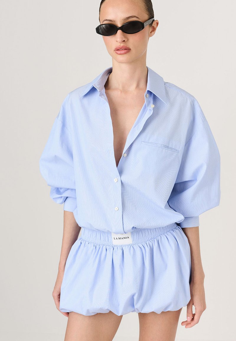Cămașă oversized de culoare albastră deschisă, cu mâneci răsucite și buzunar, asortată cu pantaloni scurți structurați lejer, având talie elastică.