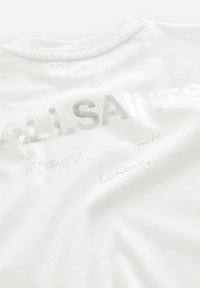 Balta medvilninė marškinėlių T-shirt su apvaliu iškirpimu. Puošti metalizuotais sidabriniais užrašais „ALLSAINTS“, „LONDON“ ir kitais tekstais įvairiomis kalbomis.