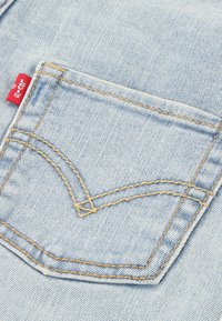 Tissu en denim bleu clair avec une poche arrière présentant une couture orange et une petite étiquette de marque rouge en tissu sur le côté gauche.