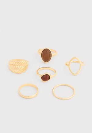 Seis anillos de oro exhibidos sobre una superficie blanca, que incluyen dos con piedras ovaladas marrones, uno texturizado, uno con forma de marquesa abierta y dos bandas lisas.