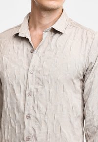 Camicia beige con bottoni, caratterizzata da un motivo testurizzato con forme geometriche, colletto e bottoni marrone chiaro. Il tessuto sembra leggero e spiegazzato.