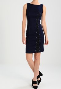 Ärmlös marinblå klänning med svart snörningsdetalj framtill, bärs med svarta blockklackskor, visas på en stående kvinna.