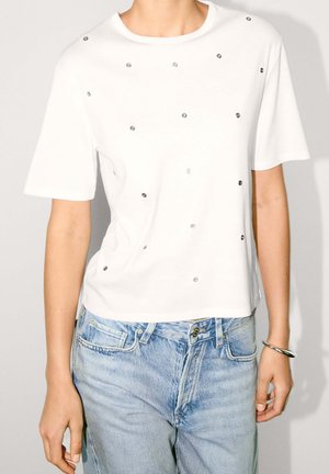 White short-sleeve t-shirt featuring metallic studs in various sizes evenly spaced across the front, paired with light blue denim jeans.