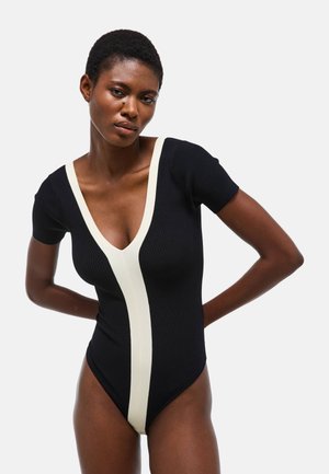 Femme aux cheveux courts portant un body noir avec un profond décolleté en V et une bande blanche verticale à l'avant, posant avec les mains dans le dos.