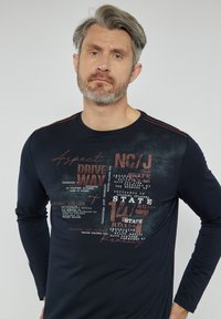 Granatowa koszula z długim rękawem z teksturowanymi grafikami w kolorze czerwonym i białym, z okrągłym dekoltem i kontrastującymi detalami szwów na ramionach.