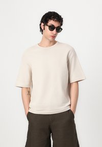 Beige oversized tričko s krátkými rukávy a kulatým výstřihem, v kombinaci s tmavými lněnými šortkami. Hladká textura, volný střih.