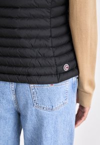 Črn napihnjen jopič ima prešito zasnovo in okrogel logotip na robu. Nosimo ga čez bež pulover in svetlo modre kavbojke.