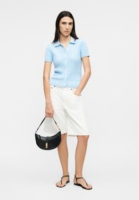 Nő, aki világoskék, rövid ujjú, gombos pulóvert és fehér, térdig érő rövidnadrágot visel, fekete kézitáskát tart a kezében, és fekete szandált visel.