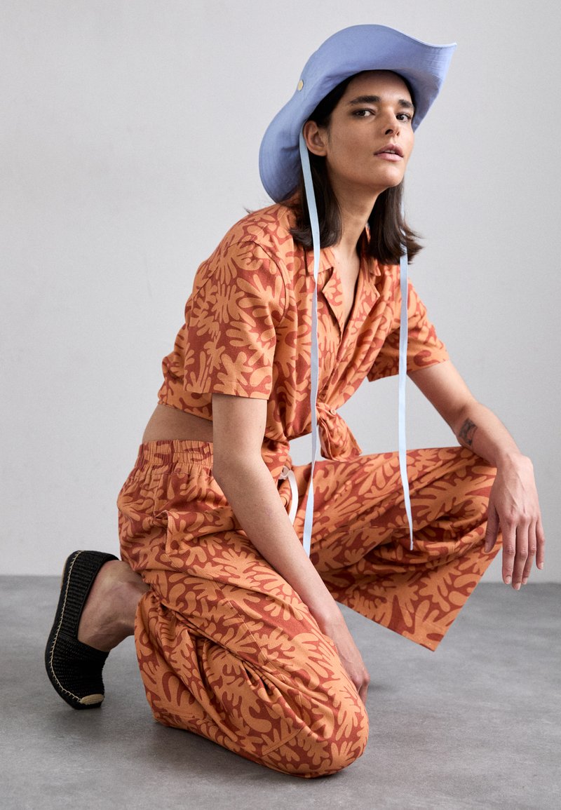 Short-sleeve orange top with a tie front and floral pattern, paired with matching loose-fitting pants. A light blue bucket hat completes the look.