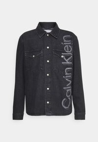 Camisa de denim negra con cierres de botones, dos bolsillos en el pecho y una gran impresión de "Calvin Klein" en un lado. Presenta un cuello clásico y mangas largas.