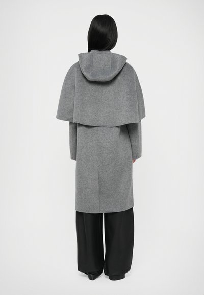 Casaco de lã cinzento com capuz, apresentando um design em camadas. O casaco é de comprimento até ao joelho e está combinado com calças largas pretas. Textura suave.