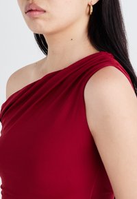 Abito rosso a spalla singola realizzato in tessuto liscio, con dettagli drappeggiati e una silhouette aderente sulla spalla di un modello.