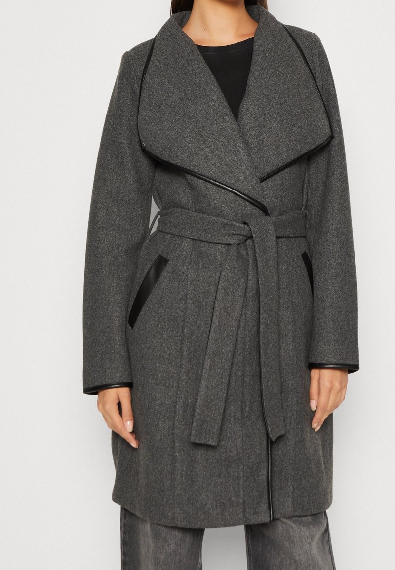 Harmaa villasekoitetakki, jossa leveä kaulus, vyötäröllä vyö, mustat nahkakoristeet taskuissa ja pitkät hihat. Sileä pinta ja istuva leikkaus.