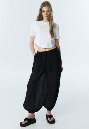Jeune femme portant un top blanc court à manches courtes, un pantalon noir ample avec des poignets élastiques et des sandales à plateforme noires, debout les mains dans les poches.