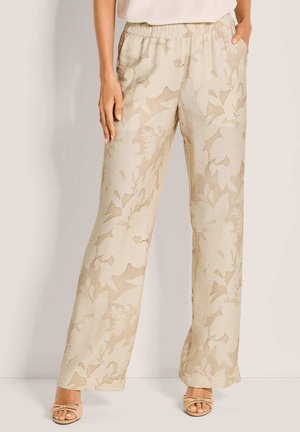 Vrouw die beige wijde broek met subtiel bloemmotief draagt, gecombineerd met naakte open teen hakken, staand tegen een effen achtergrond.