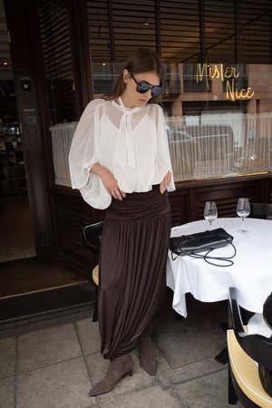 Bela, lepršava bluzica s vezirom oko vrata, uparena s smeškom, drapiranom suknjom. Smešaste čizme do članaka i crna torba na stolu.