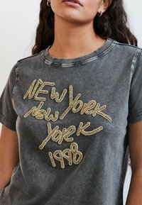 Pelēka kokvilnas T-kreklā ar nodilušu apdari un zelta pērlīšu tekstu, kas lasa "NEW York 1998". Apaļš kakls un īsas piedurknes.
