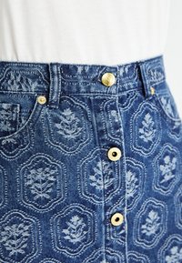 Modra denim krila z cvetličnimi vzorci, zlatimi gumbi in oprijetim pasom. Tekstura je gladka z klasičnim pet-pocket dizajnom.