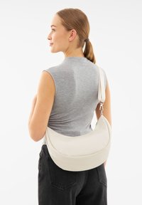 Borsa a spalla in finta pelle bianca con forma curvata, superficie liscia e tracolla regolabile, indossata in modo casual su una maglietta grigia e pantaloni neri.