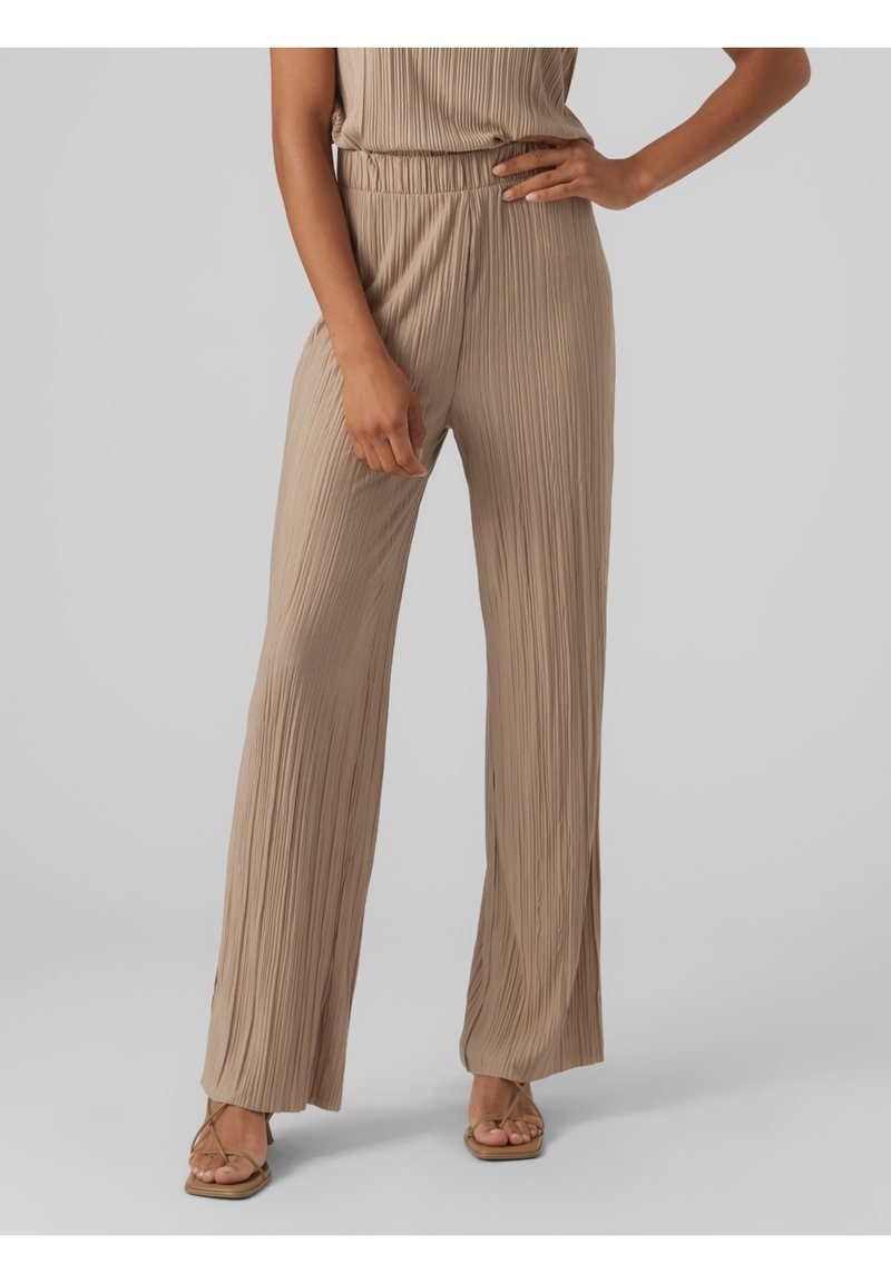 Femme portant un pantalon plissé beige à taille haute et jambes larges ainsi qu’un haut sans manches assorti, debout avec une main sur la hanche et des sandales aux pieds.