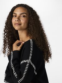Black sweater featuring white stitching accents in a crisscross pattern. Model has curly hair and a neutral background.