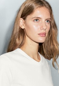 Jonge vrouw met lichtbruin haar en blauwe ogen, gekleed in een witte V-hals T-shirt, die iets naar rechts kijkt tegen een effen grijze achtergrond.