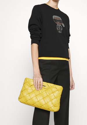 Persona que lleva una sudadera negra con un dibujo de caricatura, pantalones negros y sostiene un bolso de mano amarillo tejido frente a un fondo blanco liso.