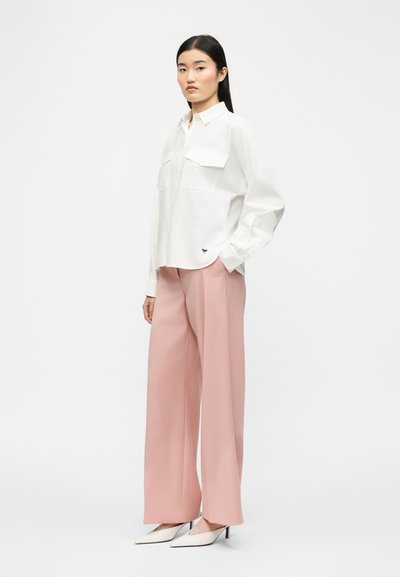 Γυναίκα που στέκεται πλαγιαστά φορώντας ένα λευκό πουκάμισο με κουμπιά και τσέπες, φαρδιά ροζ παντελόνια και λευκά μυτερά ψηλοτάκουνα παπούτσια.