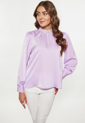 Frau mit langen, welligen braunen Haaren, die eine glänzende hellviolette Bluse mit Puffärmeln und eine weiße Hose vor einem einfarbigen weißen Hintergrund trägt.