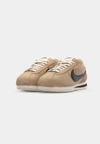 Kreminės sportinės bateliai su lygaus nubuko viršumi, juodu Nike swoosh ženkliuku, baltais raišteliais ir tekstūruota padu. Minimalistinis dizainas, klasikinė siluetas.