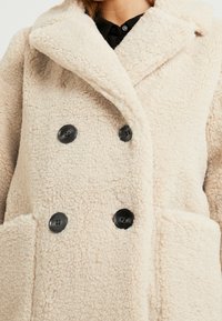 Abrigo de teddy color crema con grandes botones negros, amplio cuello y bolsillos de parche, llevado sobre una camisa negra con cuello.