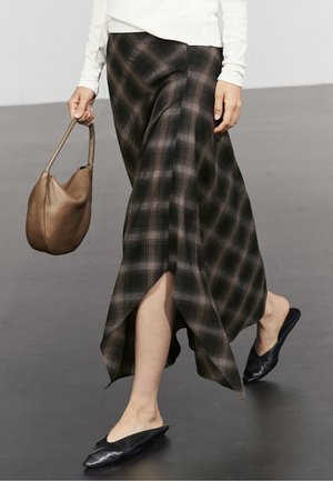 Persona che cammina indossando una gonna asimmetrica a quadri nera e marrone, ballerine nere con punta, una maglia bianca a manica lunga e che tiene una borsa marrone.