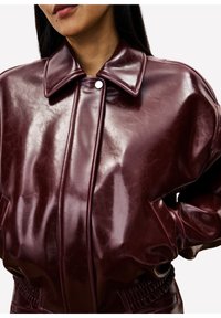 Vrouw in een glanzende donker bordeaux lederen jas met een brede kraag en zijzakken, met handen op de heupen, kort bij de nek en taille.