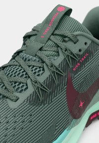 Primo piano di una scarpa da corsa Nike Pegasus Trail 5 verde con swoosh bordeaux e lettering rosa su tessuto in rete e suola testurizzata.