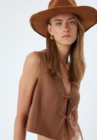 Top marrone senza maniche con scollatura a V, caratterizzato da due legature frontali. Tessuto morbido con una vestibilità rilassata. Indossato con un cappello marrone a tesa larga.
