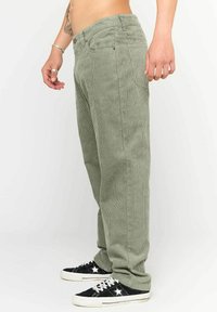 Pantaloni di velluto a coste verde con righe verticali, cinque tasche e chiusura con bottone. Abbinati a sneaker nere con dettagli bianchi.