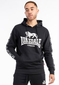 Black hoodie made of cotton blend, featuring a large white lion logo and "Lonsdale London" text on the front. Front kangaroo pocket and drawstring.