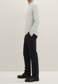 Maglione grigio chiaro lavorato a maglia con scollatura a V e polsini a coste, abbinato a pantaloni blu navy scuri e scarpe nere stringate, mostrato di profilo.