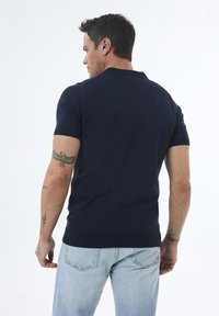 Camisa polo de manga corta en color navy, con un diseño ajustado, cuello y dobladillo acanalados, combinada con jeans de color azul claro. Presenta tatuajes visibles.