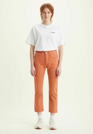 T-shirt bianco in cotone con logo nero, abbinato a pantaloni cropped in denim arancione, con design a gamba dritta e tasche frontali. Sneakers bianche.
