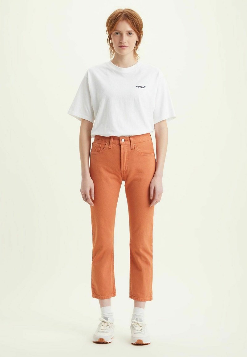 T-shirt bianco in cotone con logo nero, abbinato a pantaloni cropped in denim arancione, con design a gamba dritta e tasche frontali. Sneakers bianche.