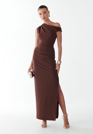 Kobieta w brązowej sukni na jedno ramię z rozcięciem do uda, trzymająca małą kopertówkę, złotą bransoletkę, kolczyki i sandały na cienkich paskach.