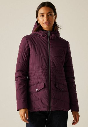 Kobieta ubrana w fioletową pikowaną kurtkę z kapturem, zamkiem z przodu, kieszeniami na klapki oraz ciemnymi spodniami, stojąca na jednolitym tle.